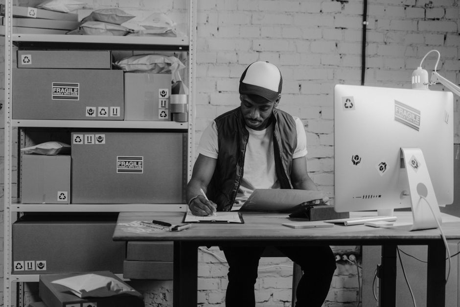Warehouse worker organizing shipments at a desk, surrounded by packages and a computer, illustrating logistics work.